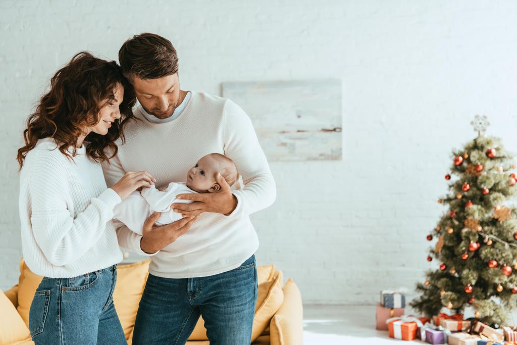 Glückliche Eltern stehen neben geschmücktem Weihnachtsbaum und halten entzückendes Baby in der Hand - Foto, Bild
