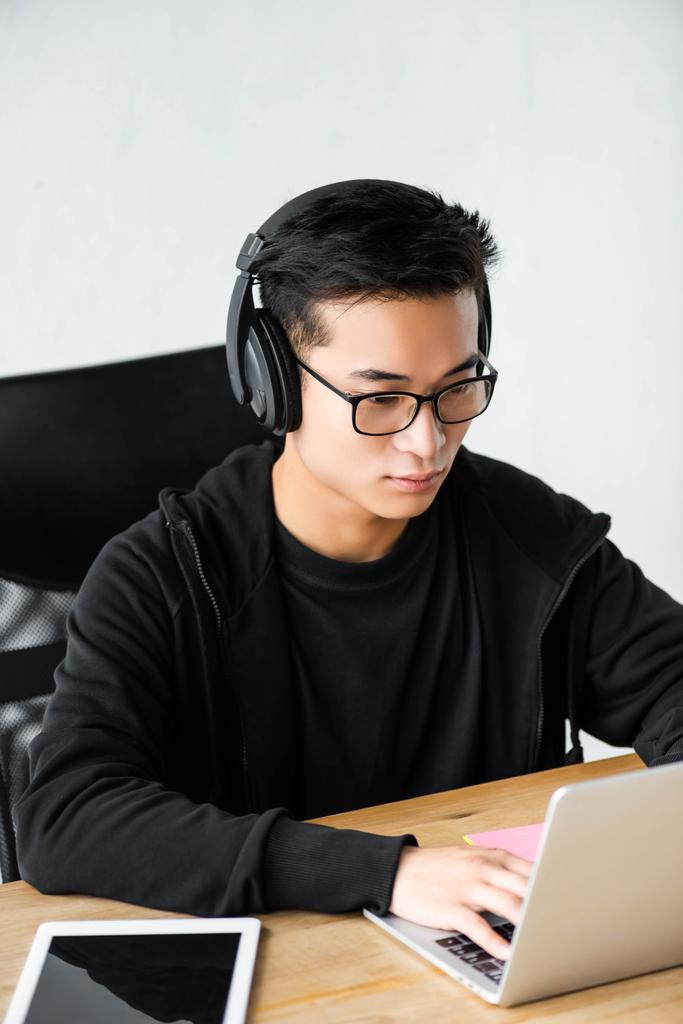 asian hacker in headphones using laptop and sitting at table  - Photo, Image