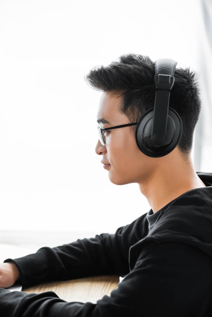 Seitenansicht eines asiatischen Hackers mit Kopfhörer am Tisch  - Foto, Bild
