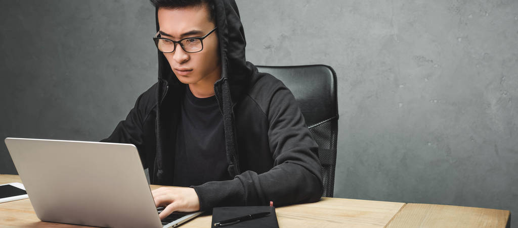 πανοραμική λήψη του ασιατικού hacker χρησιμοποιώντας φορητό υπολογιστή και κάθεται στο τραπέζι  - Φωτογραφία, εικόνα