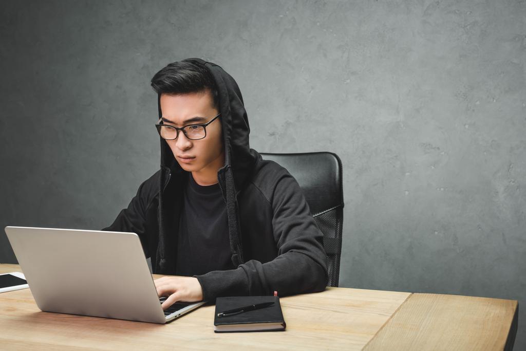 Gözlüklü Asyalı hacker dizüstü bilgisayar kullanıyor ve masada oturuyor.  - Fotoğraf, Görsel