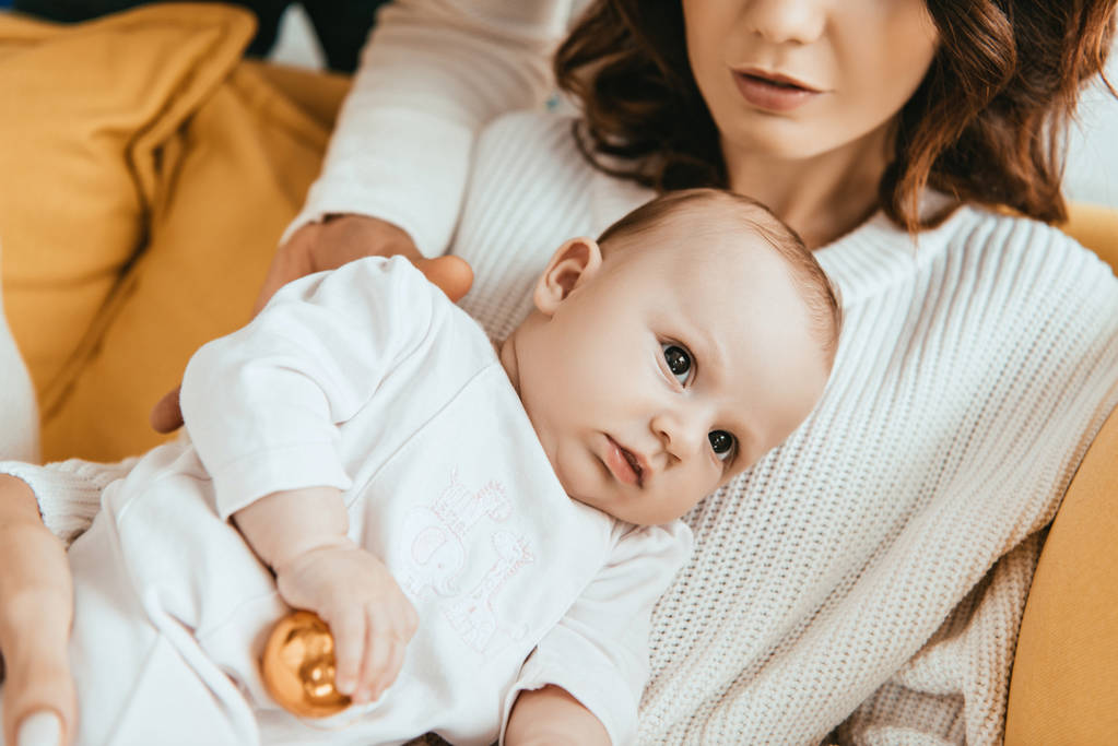 vista recortada de la mujer sentada en el sofá con adorable bebé sosteniendo la bola de Navidad
 - Foto, imagen
