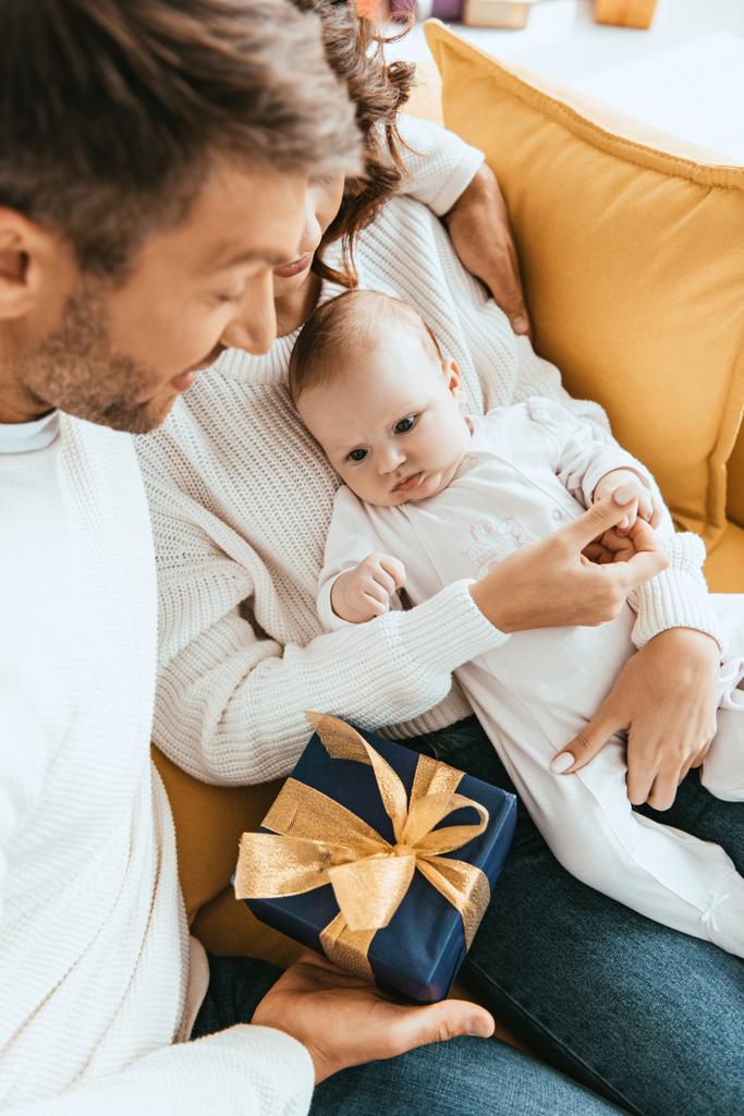 Vater überreicht Geschenkbox an entzückende kleine Tochter, die auf den Händen der Mutter sitzt - Foto, Bild