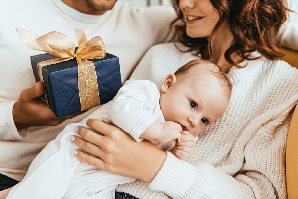 cropped view of husband presenting gift box to happy wife holding adorable baby - Photo, Image