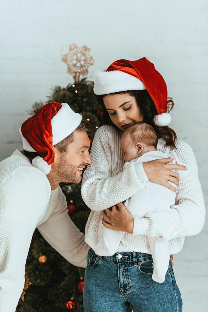 attraktive Frau mit Weihnachtsmannmütze mit entzückendem Baby in der Nähe eines glücklichen Ehemanns - Foto, Bild