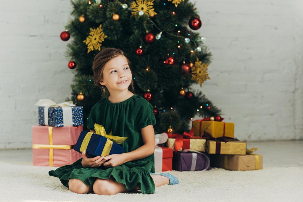 щаслива дитина сидить на підлозі біля ялинки, тримає подарункову коробку і дивиться
 - Фото, зображення