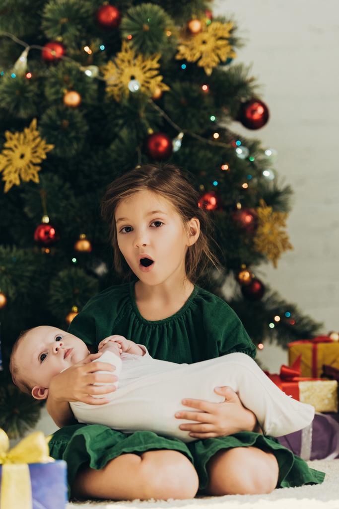 amüsiertes Kind blickt in die Kamera, während es neben dem Weihnachtsbaum sitzt und seine süße kleine Schwester hält - Foto, Bild