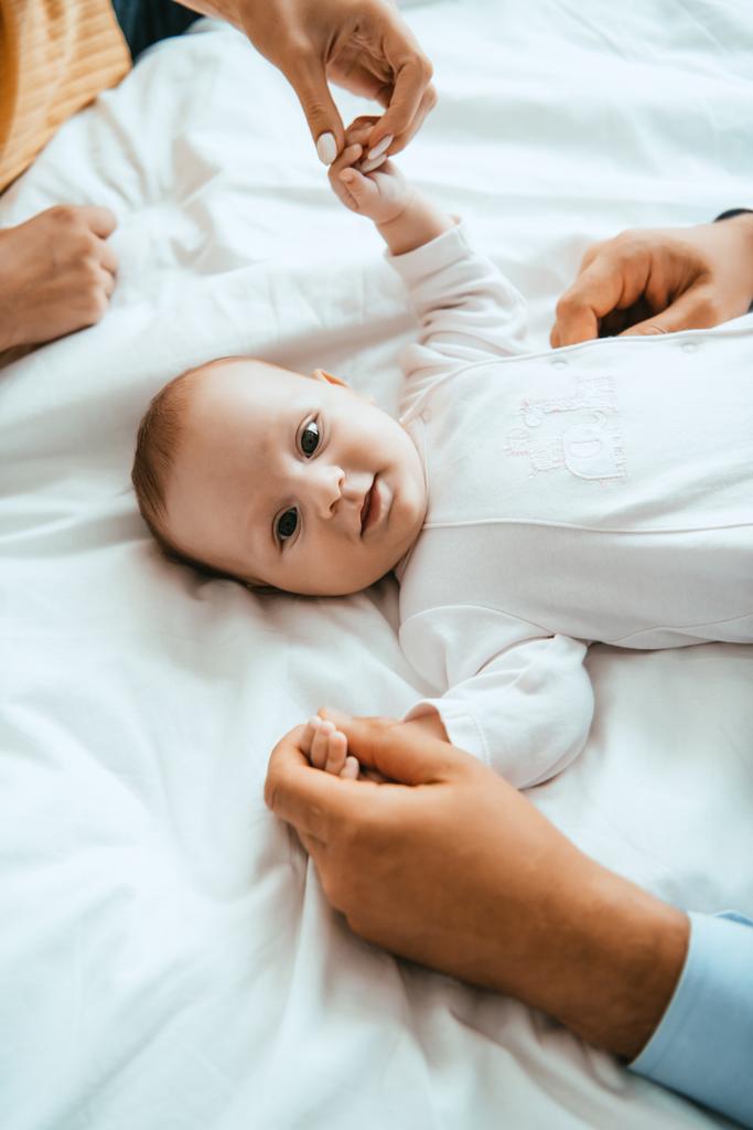 κορυφαία άποψη της μητέρας και του μπαμπά παίζει με χαμογελαστό μωρό που βρίσκεται σε λευκό κρεβάτι - Φωτογραφία, εικόνα