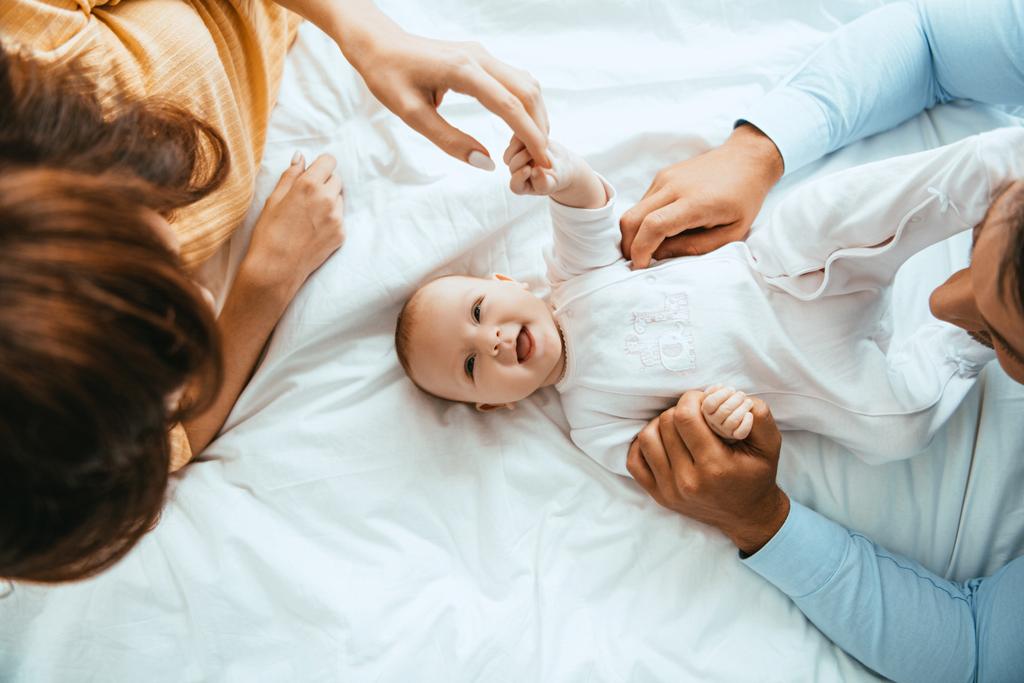 widok rodziców dotykających uśmiechniętego niemowlęcia leżącego na białej pościeli - Zdjęcie, obraz