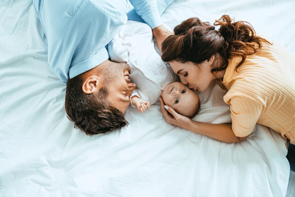 счастливая мать целует ребенка, лежа на кровати рядом с улыбающимся мужем
 - Фото, изображение