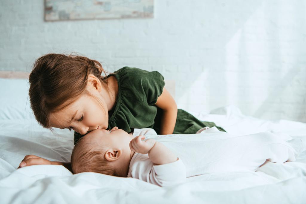 Küçük kız kardeşini öpen sevimli çocuk beyaz yatağa uzanıyor. - Fotoğraf, Görsel