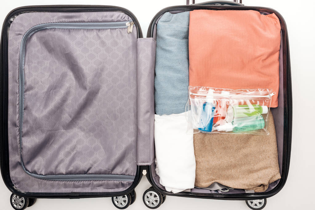 Havlulu seyahat çantasının üst görüntüsü, şişeli kozmetik çanta, beyaz arka planda giysiler.  - Fotoğraf, Görsel