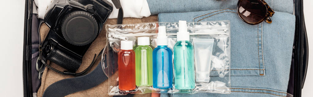 panoramatický záběr cestovní tašky s ručníkem, kosmetickou taškou s barevnými láhvemi, digitálním fotoaparátem, oblečením a slunečními brýlemi - Fotografie, Obrázek