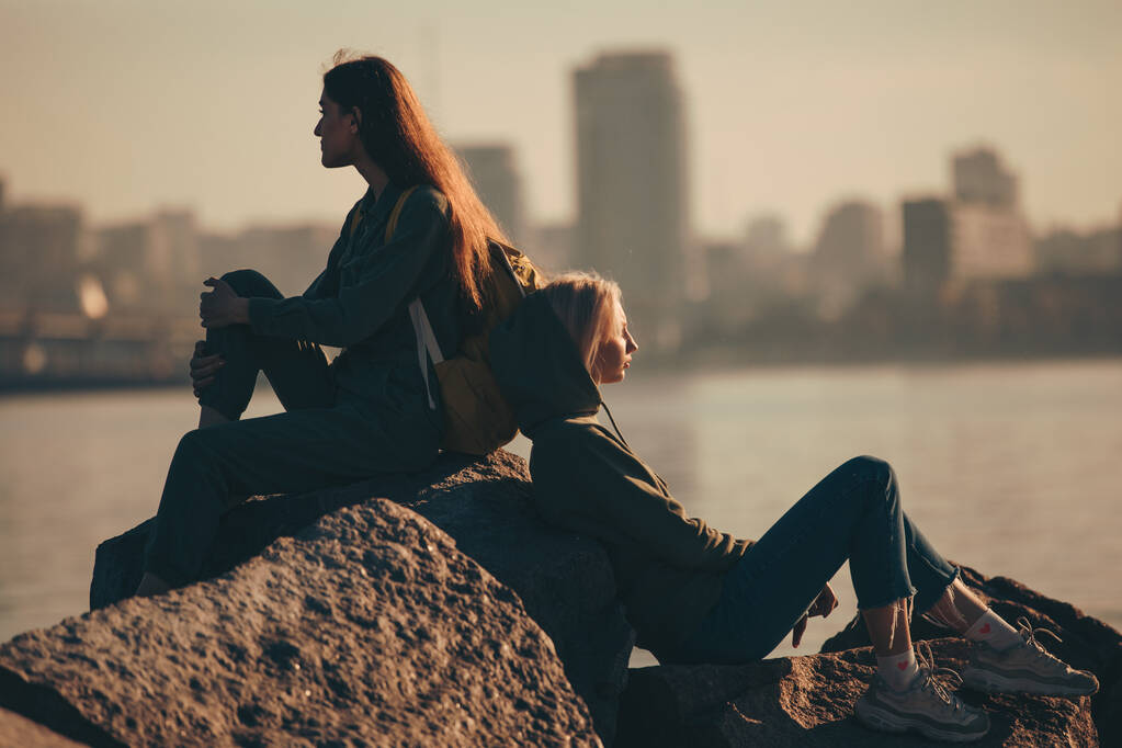 Silhueta de duas jovens mulheres sentadas em pedras perto do rio
 - Foto, Imagem