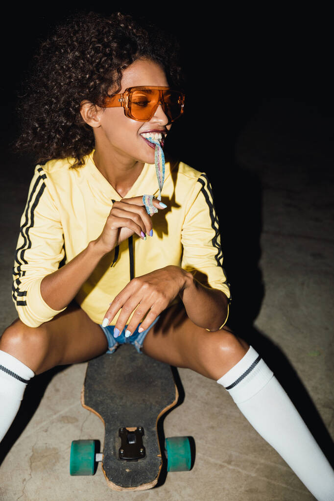 Obrázek africké americké dívky jíst dlouhé cukroví na skateboardu a - Fotografie, Obrázek