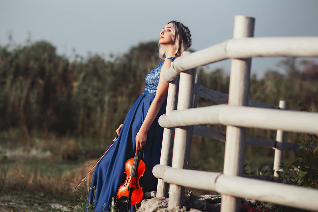 portrait of a beautiful girl in a long dress with a violin near a wooden painted fence, a young woman walks with a musical instrument outdoors - Photo, Image
