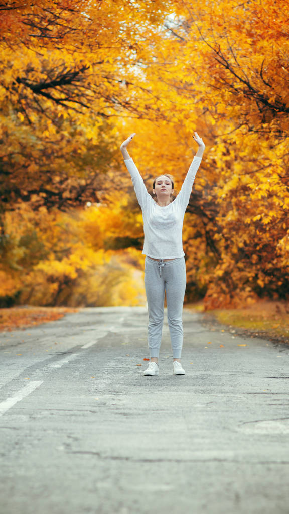 スポーツウェアの練習でスリムな若い女性とアスファルトの道路に立ち上がる手を伸ばし、女の子は秋の葉を背景に屋外スポーツに従事し、コンセプト健康的なライフスタイルと女性の美しさ - 写真・画像