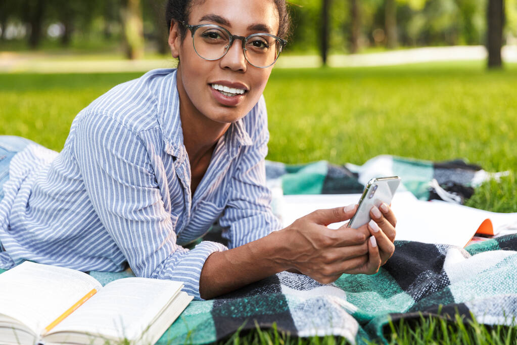公園でスマートフォンを持っている幸せなアフリカ系アメリカ人女性のイメージ - 写真・画像