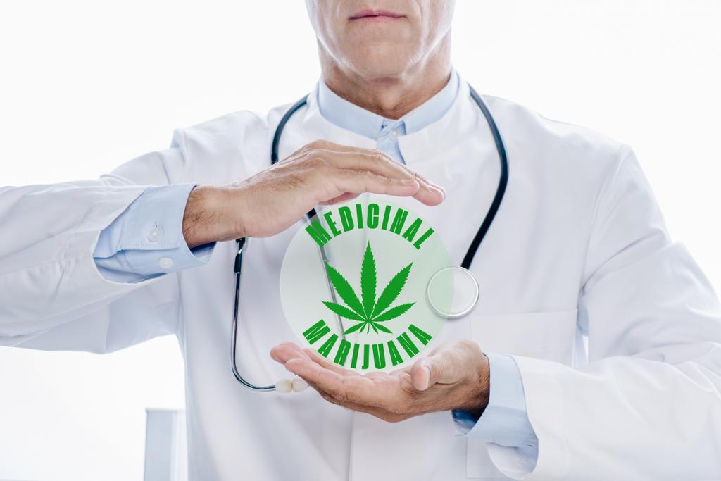 cropped view of doctor in white coat holding hands around medicinal marijuana illustration isolated on white - Photo, Image