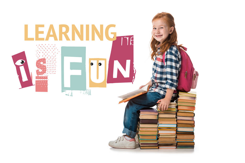 cheerful redhead pupil smiling while sitting on books near learning is fun letters on white  - 写真・画像