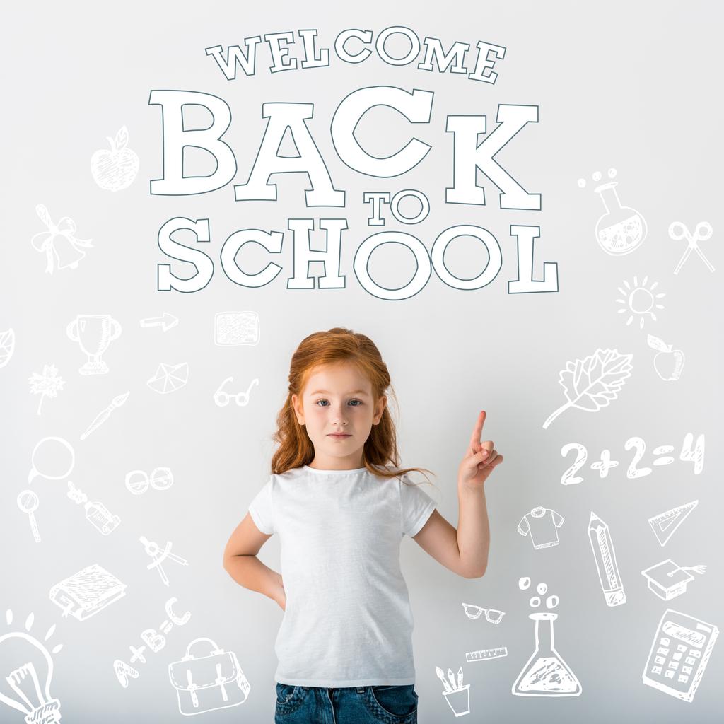 niedliche rothaarige Kind blickt in die Kamera und zeigt mit dem Finger in der Nähe Willkommen zurück zur Schule Buchstaben auf weiß  - Foto, Bild