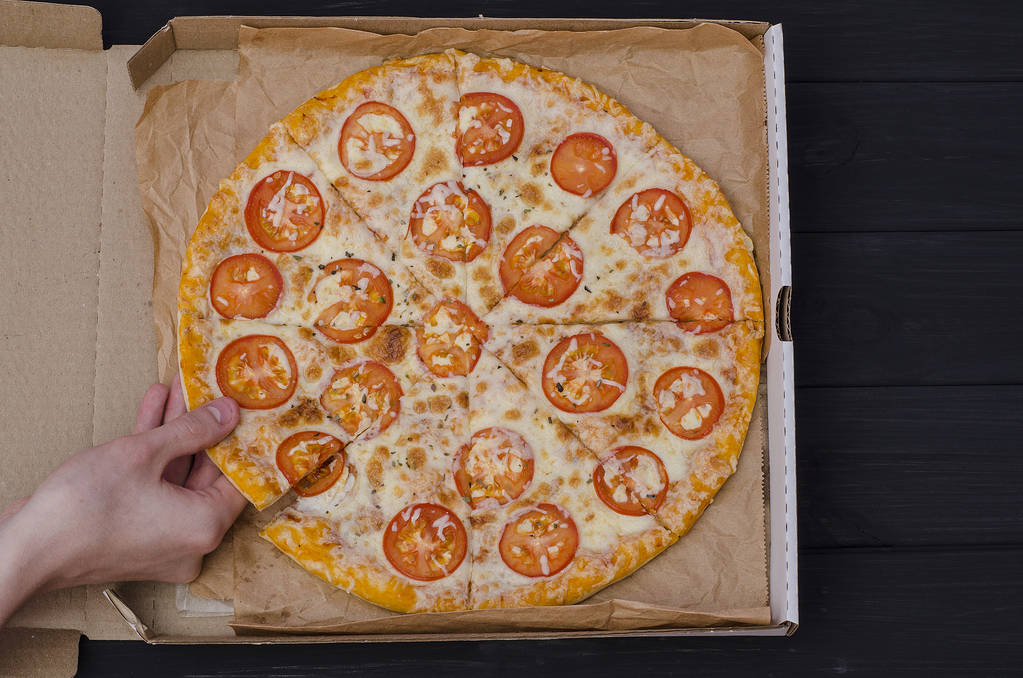 Pizza con queso y tomates en una caja sobre fondo de madera negra. Un joven toma una rebanada de pizza. Pizzerías publicitarias
 - Foto, Imagen