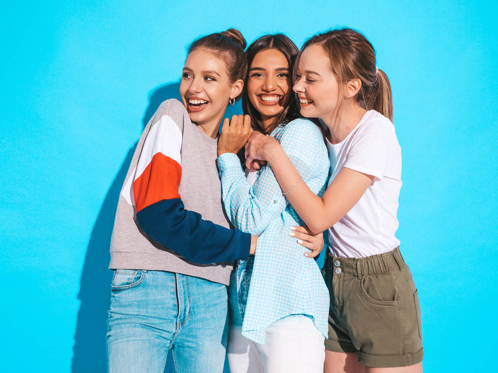 Três jovens bonitas meninas hipster sorridentes em roupas de verão na moda. Mulheres despreocupadas Sexy posando perto da parede azul no estúdio. Modelos positivos se divertindo e abraçando
 - Foto, Imagem