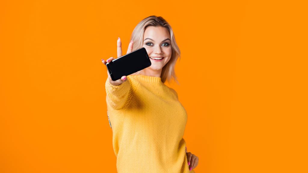 młoda blondynka w żółtym swetrze trzyma smartfona w ręce z pustym ekranem, makieta. - Zdjęcie, obraz