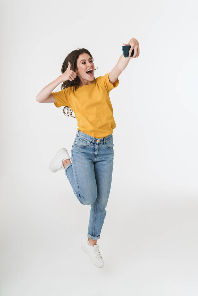 Optymistyczna młoda emocjonalna kobieta skacze odizolowany na białym tle ściany zrobić selfie przez telefon komórkowy. - Zdjęcie, obraz