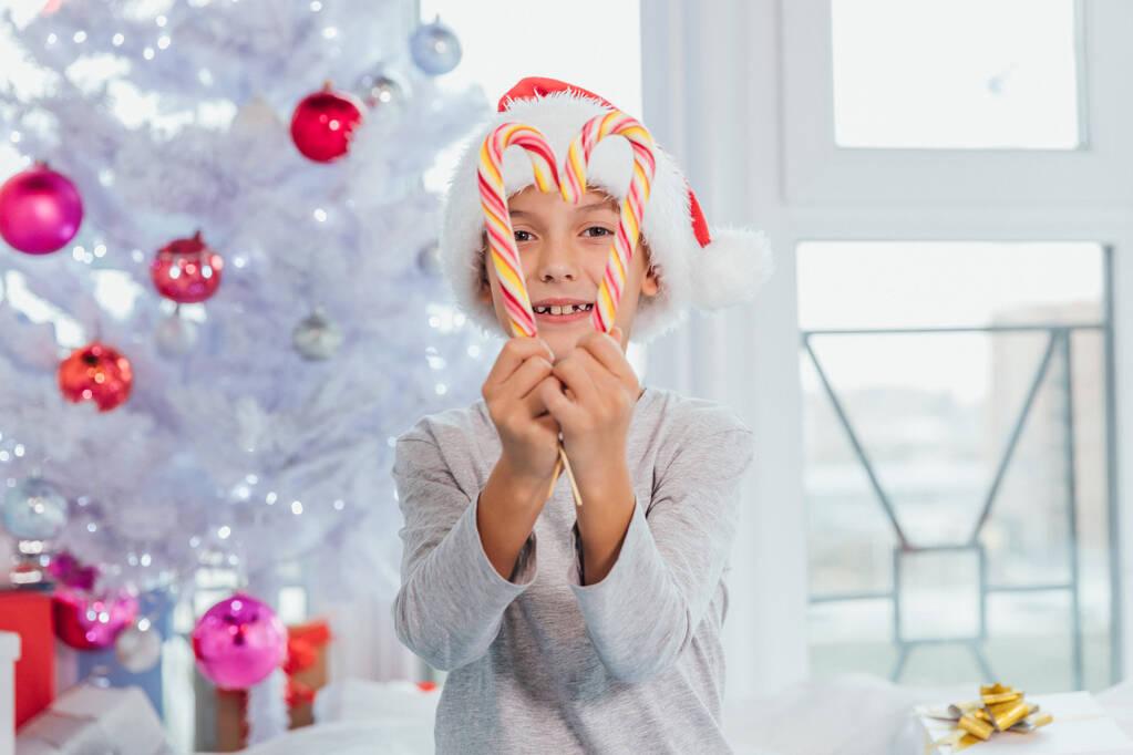 Portré boldog kiskorú fiú piros Mikulás kalap néz át a szíve karácsonyi cukornád, kezében édességet az arca előtt. - Fotó, kép