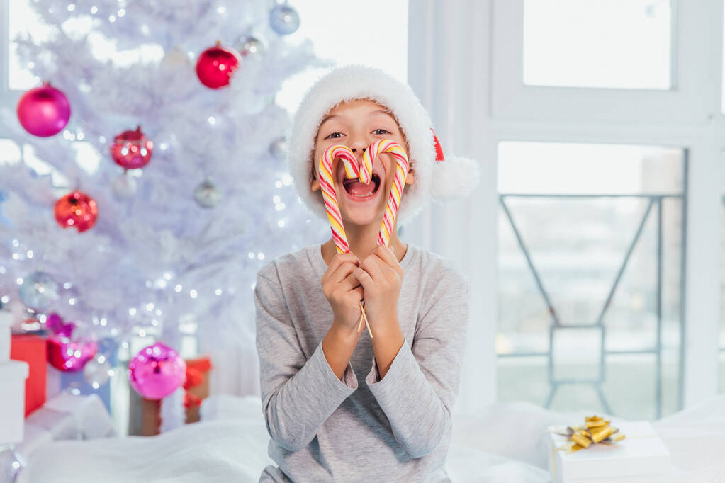 Kleine zoetekauw jongen houdt heerlijke snoepjes in de voorkant van de mond, kijkt direct naar de camera, lachen, geïsoleerd over witte nieuwjaar achtergrond. - Foto, afbeelding