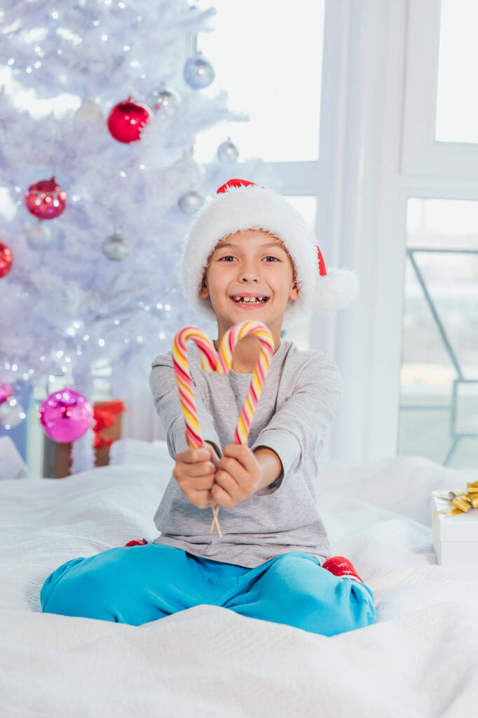 Noel Baba şapkalı komik çocuk pijamalarıyla yatakta oturmuş çizgili şeker kamışlarını gösteriyor.. - Fotoğraf, Görsel