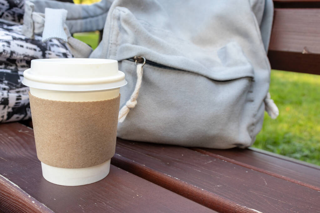 Jednorazowa papierowa filiżanka kawy i plecak na ławce w parku, hamulec kawowy na świeżym powietrzu - Zdjęcie, obraz