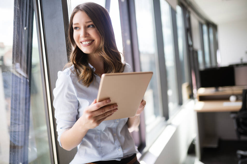 Jeune femme d'affaires utilisant une tablette numérique tout en se tenant devant les fenêtres dans le bureau
 - Photo, image