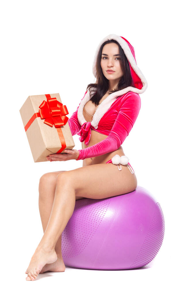 Natale. Sport fitness girl in un costume rosso sexy Babbo Natale in possesso di una scatola regalo legato con un nastro rosso con un fiocco e seduto su una palla fitness. Isolato su sfondo bianco
. - Foto, immagini