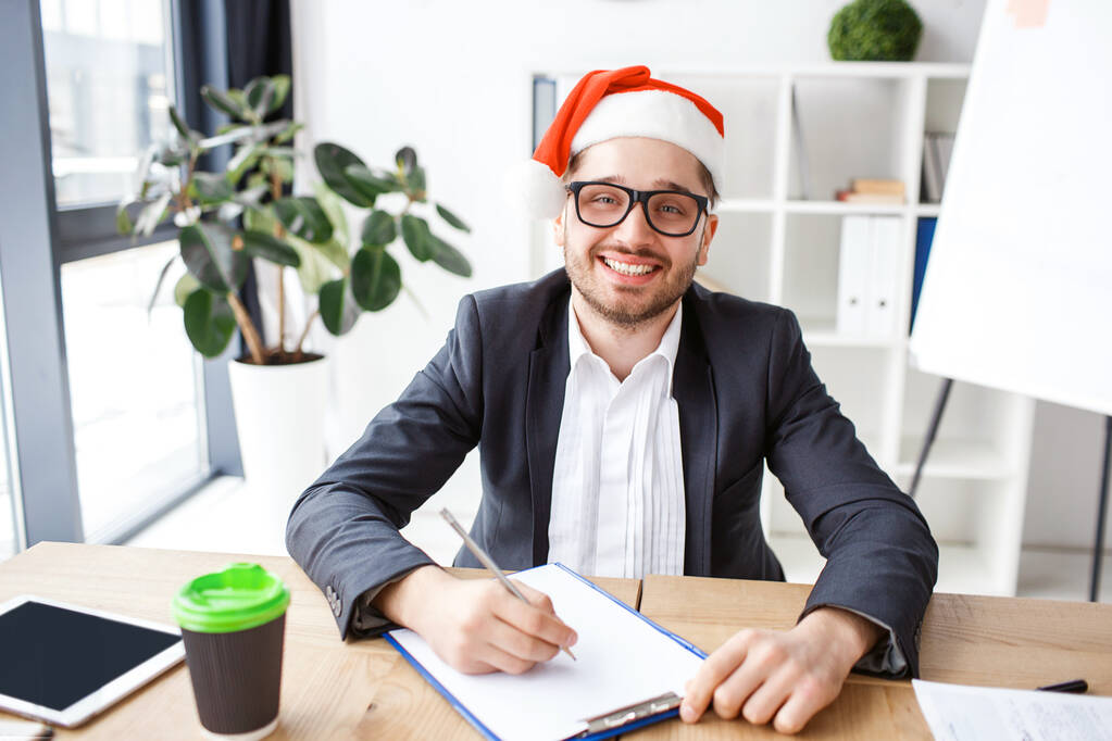 Νεαρός χαρούμενος επιχειρηματίας στο γραφείο. Γιορτάζουμε την Πρωτοχρονιά ή τα Χριστούγεννα. Να δουλεύω μόνος. Γράφω σημειώσεις. Γιορτινή διάθεση. Θετικά χαρούμενος καλός άνθρωπος. - Φωτογραφία, εικόνα