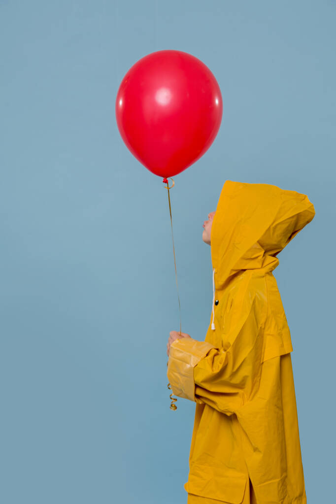 Mały chłopiec w żółtym płaszczu przeciwdeszczowym z czerwonym balonem na niebieskim tle - Zdjęcie, obraz