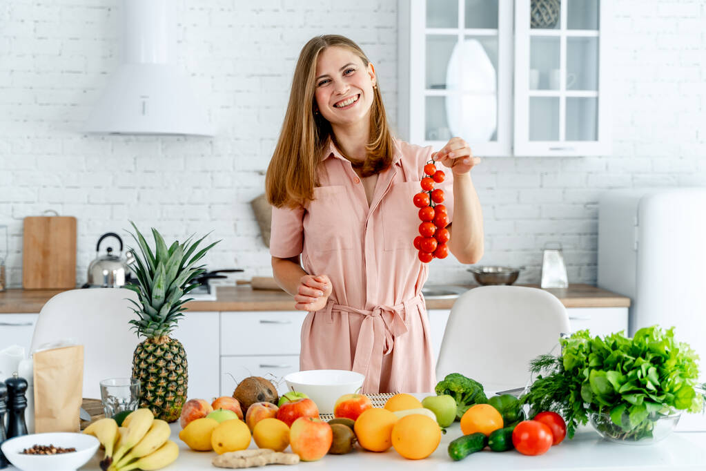 Vrouw in de keuken klaar om te bereiden maaltijd met groenten en fruit. De vrouw houdt tomaten vast. Keuken achtergrond. Gezond eten. Veganisten. Vegetarisch. Bankje tomaten. - Foto, afbeelding
