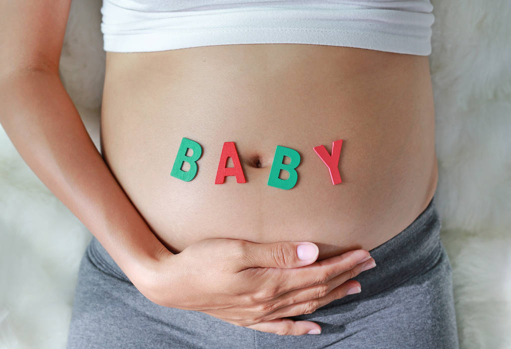 Nahaufnahme einer schwangeren Frau, die auf einem weichen Sofa sitzt und ihren Bauch mit einem Baby vor ihrem Bauch berührt. - Foto, Bild