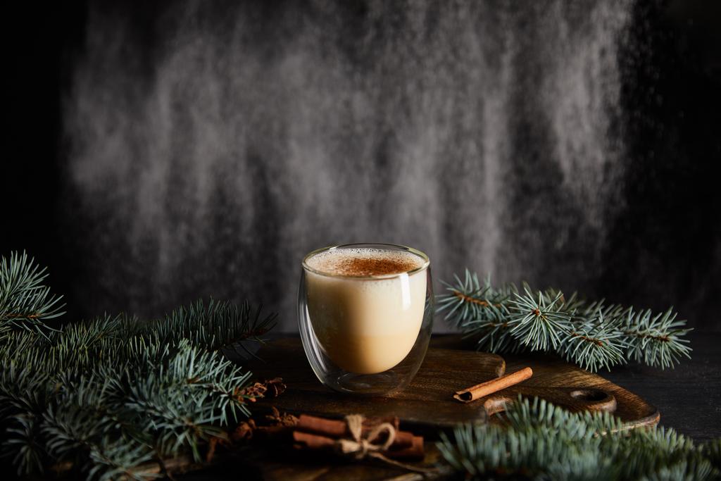 tasty eggnog cocktail on cutting board near spruce branches and cinnamon sticks on black background with powdered sugar falling like snow - Photo, Image