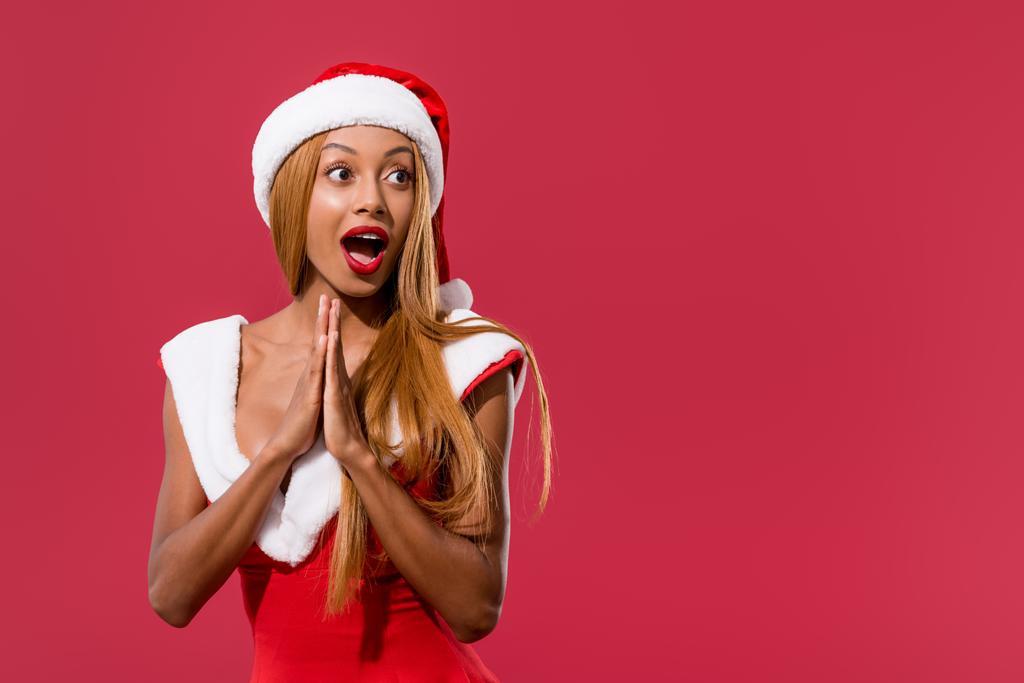 amüsiertes afrikanisch-amerikanisches Mädchen mit Weihnachtsmütze und Weihnachtskleid, das mit gefalteten Händen steht und isoliert auf rotem Grund wegschaut - Foto, Bild