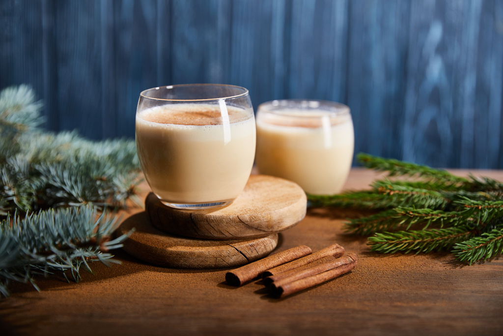tasty eggnog cocktail on round wooden boards near spruce branches and cinnamon sticks on blue textured background  - Photo, Image