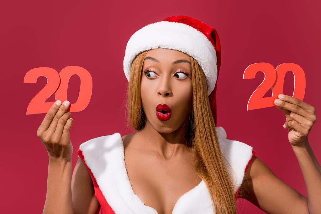 shocked african american girl in santa hat and christmas dress holding 2020 paper cut number isolated on red - 写真・画像