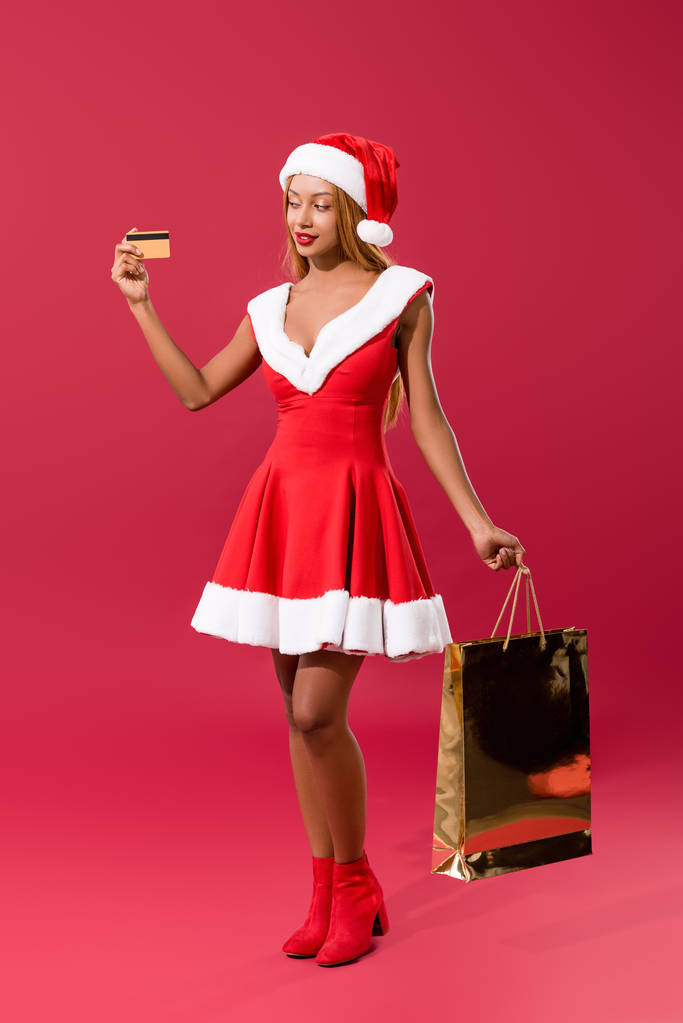 menina americana africana atraente em santa chapéu e vestido de Natal segurando cartão de crédito e saco de compras no fundo vermelho
 - Foto, Imagem