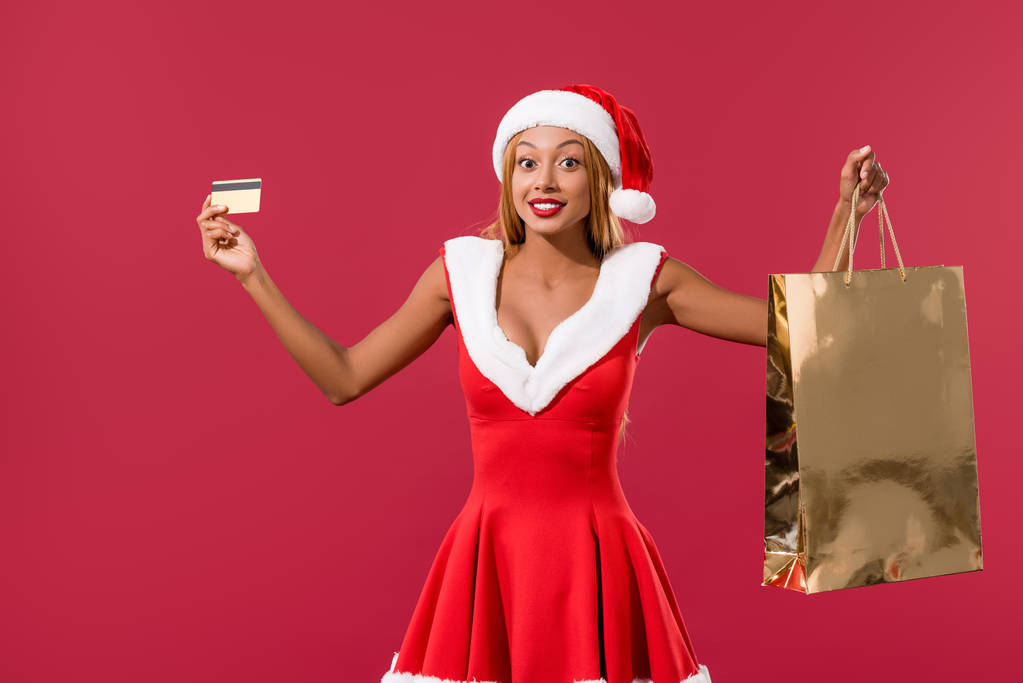 divertida chica afroamericana en sombrero de santa y vestido de navidad celebración de tarjeta de crédito y bolsa de compras aislado en rojo
 - Foto, imagen