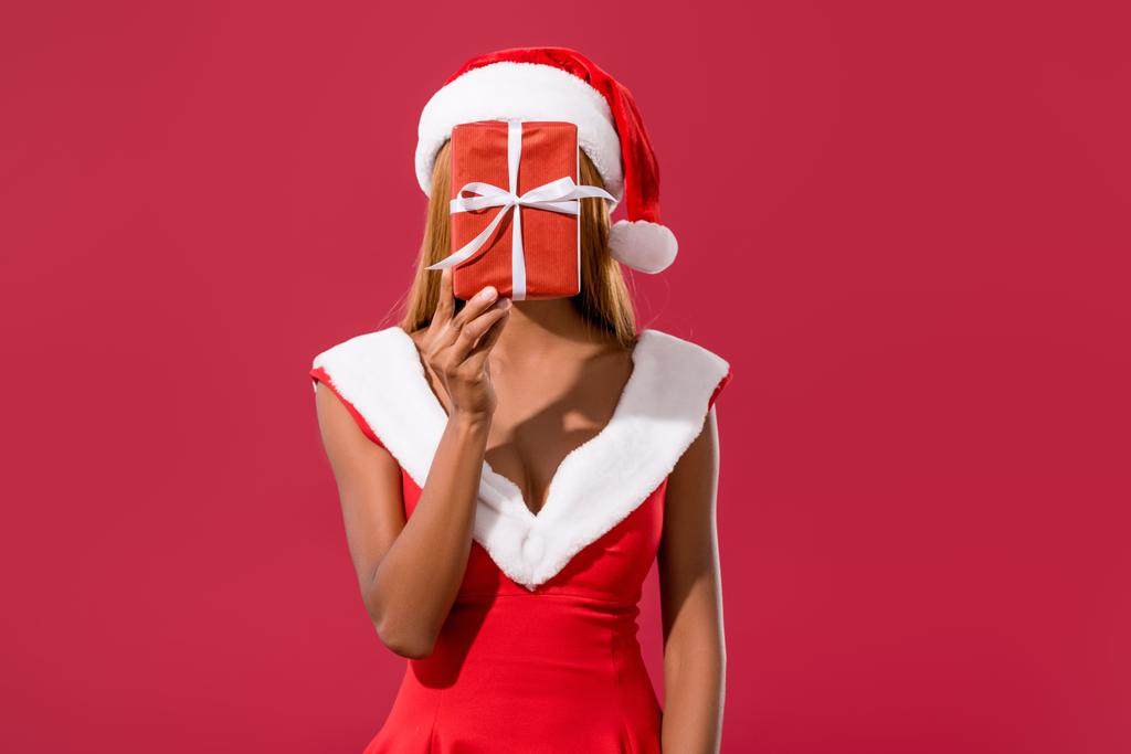 young african american girl in santa hat and christmas dress hiding face behind gift box isolated on red - Photo, Image
