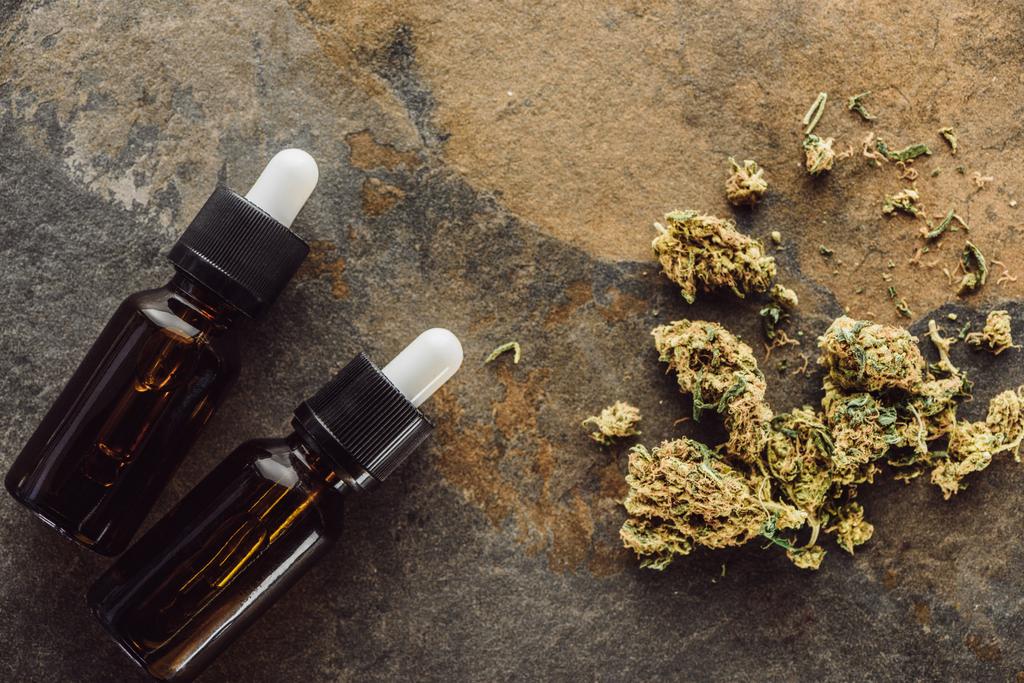 bovenaanzicht van medicinale marihuana knoppen in de buurt van flessen met hennepolie op marmeren oppervlak - Foto, afbeelding