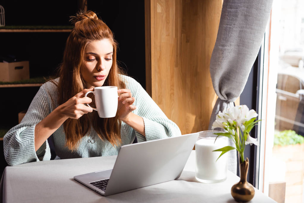 atrakcyjna ruda kobieta pije kawę i korzysta z laptopa w kawiarni - Zdjęcie, obraz