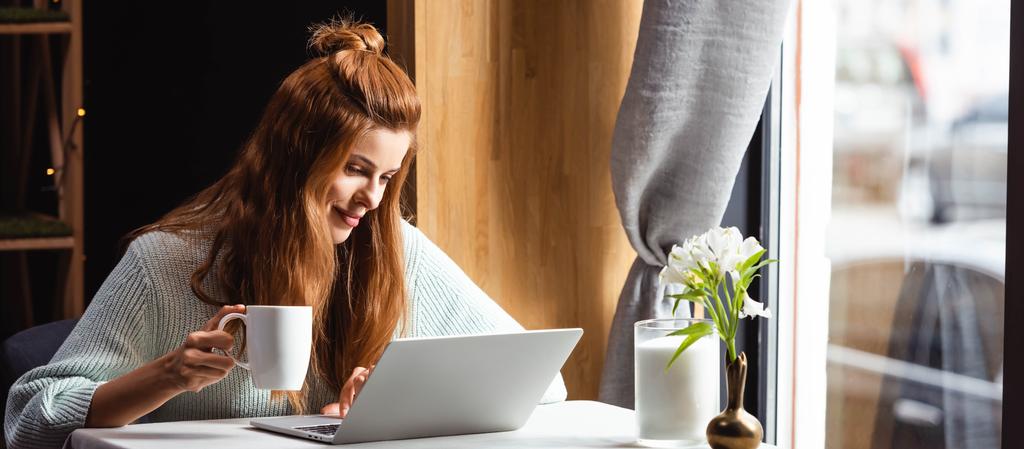 kaunis hymyilevä nainen kupin kahvia kannettavan tietokoneen avulla kahvilassa
 - Valokuva, kuva