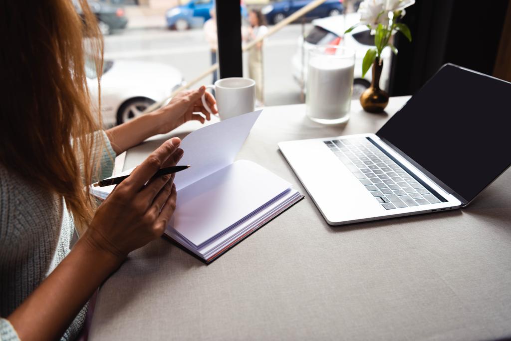обрезанный вид девушка учится онлайн с блокнотом и ноутбуком в кафе с чашкой кофе
   - Фото, изображение